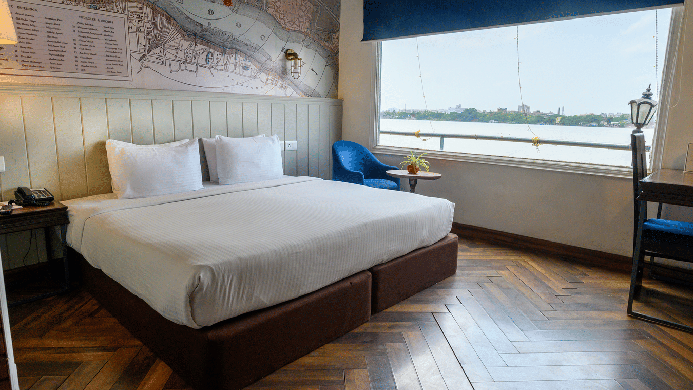 A stylish hotel bedroom featuring a bed with white bedding, a blue armchair, and a large window overlooking the water.