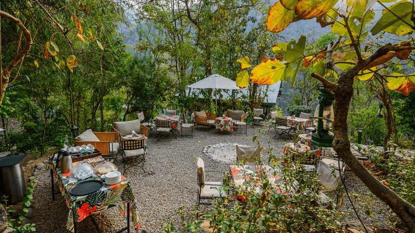 An outdoor dining area set amidst lush greenery, featuring wooden tables and a gazebo at Neemrana's Glasshouse on The Ganges.