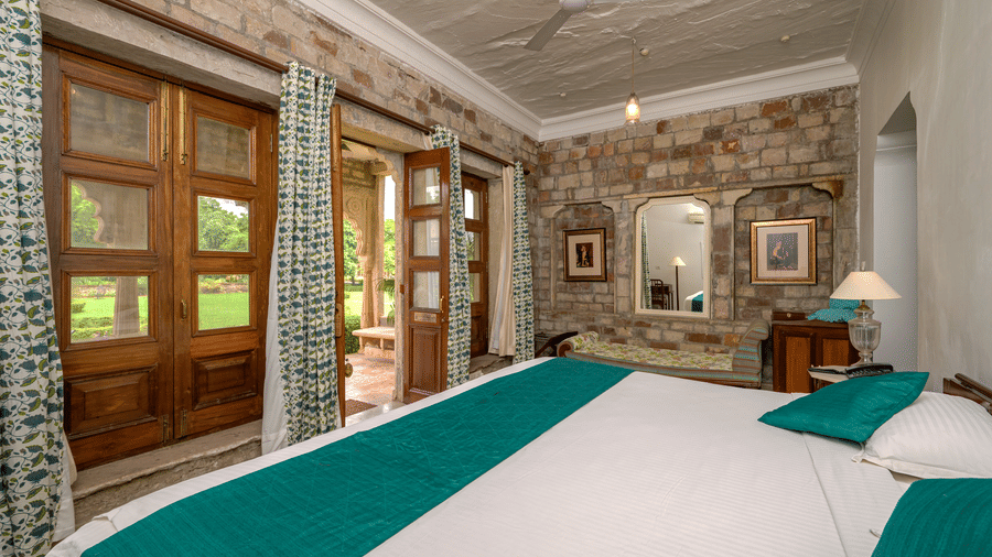Spacious heritage bedroom featuring carved wooden doors, stone walls, patterned curtains, elegant furnishings, and a large bed overlooking lush gardens at Deo Bagh, Gwalior