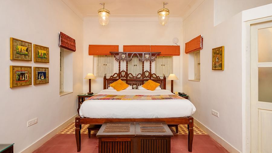 A bedroom with a wooden four-poster bed, orange accents, framed artwork on the walls, and a patterned rug, highlighting the room's traditional decor - Tijara Fort-Palace - 19th Century, Alwar.