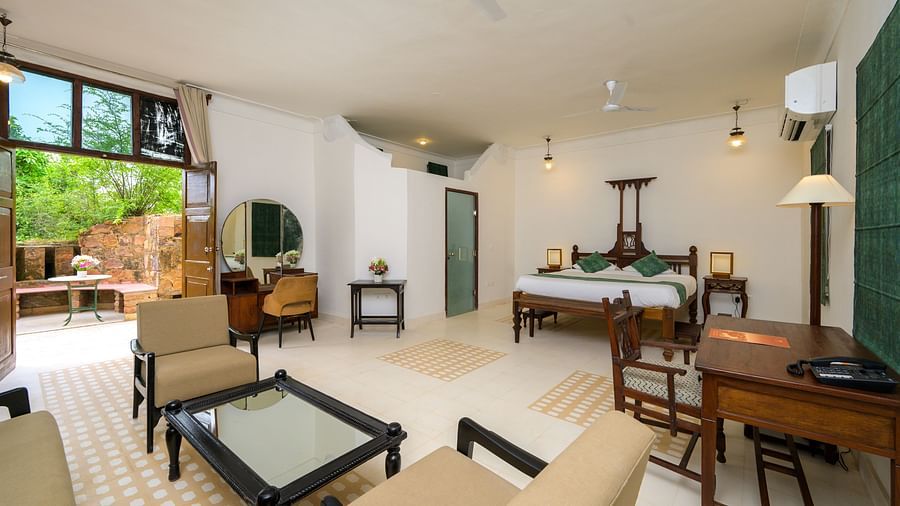 A spacious room featuring a wooden four-poster bed, a seating area with beige chairs, a dark wood desk, and an open doorway leading to an outdoor space, offering a blend of indoor and outdoor living - Tijara Fort-Palace - 19th Century, Alwar.