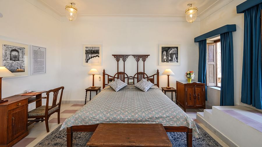 A room with a bed, a desk, a wooden bench, and two framed artworks in Tijara Fort-Palace - 19th Century, Alwar.