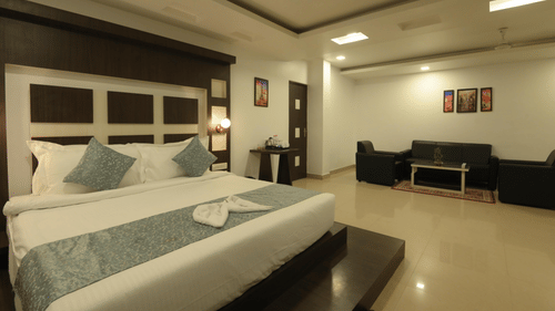 Suite at Biznotel by Pride, Deoghar featuring a plush bed with fresh linen with a large headboard and a sitting space with sofa set and coffee table in the background under warm lighting