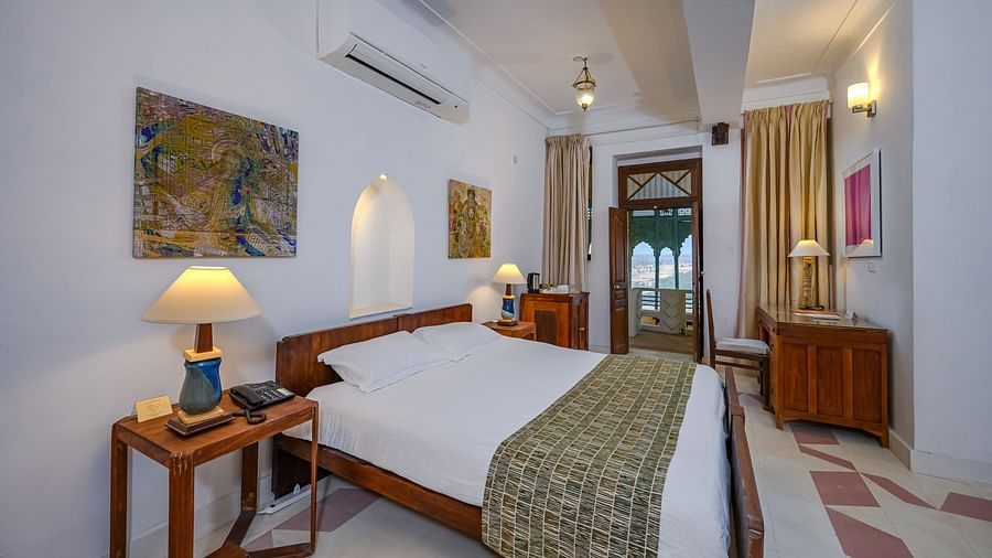 A bed with white linens and a patterned throw is in a bedroom at Tijara Fort-Palace - 19th Century, Alwar, which also features bedside tables with lamps, artwork on the walls, and a glimpse into an adjacent area.