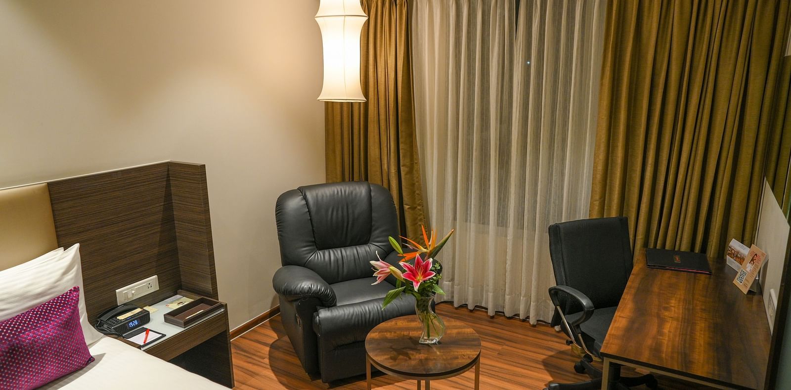 A bedroom featuring a bed, side table, work desk with chair, telephone, black armchair, overhead lamp, and a coffee table.