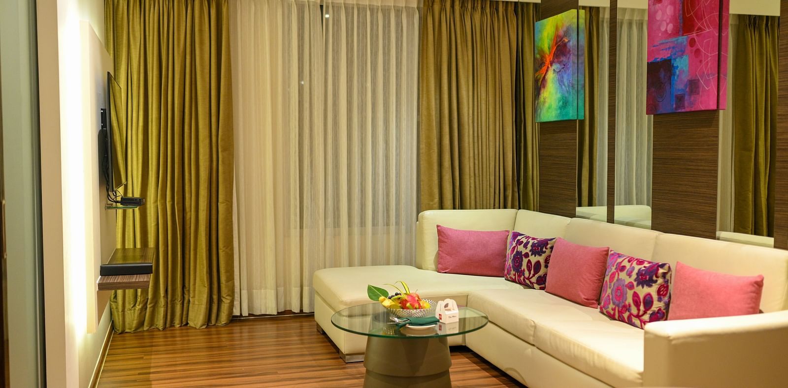 A living area featuring a couch adorned with colorful pillows, a small coffee table, wall paintings, and a TV.