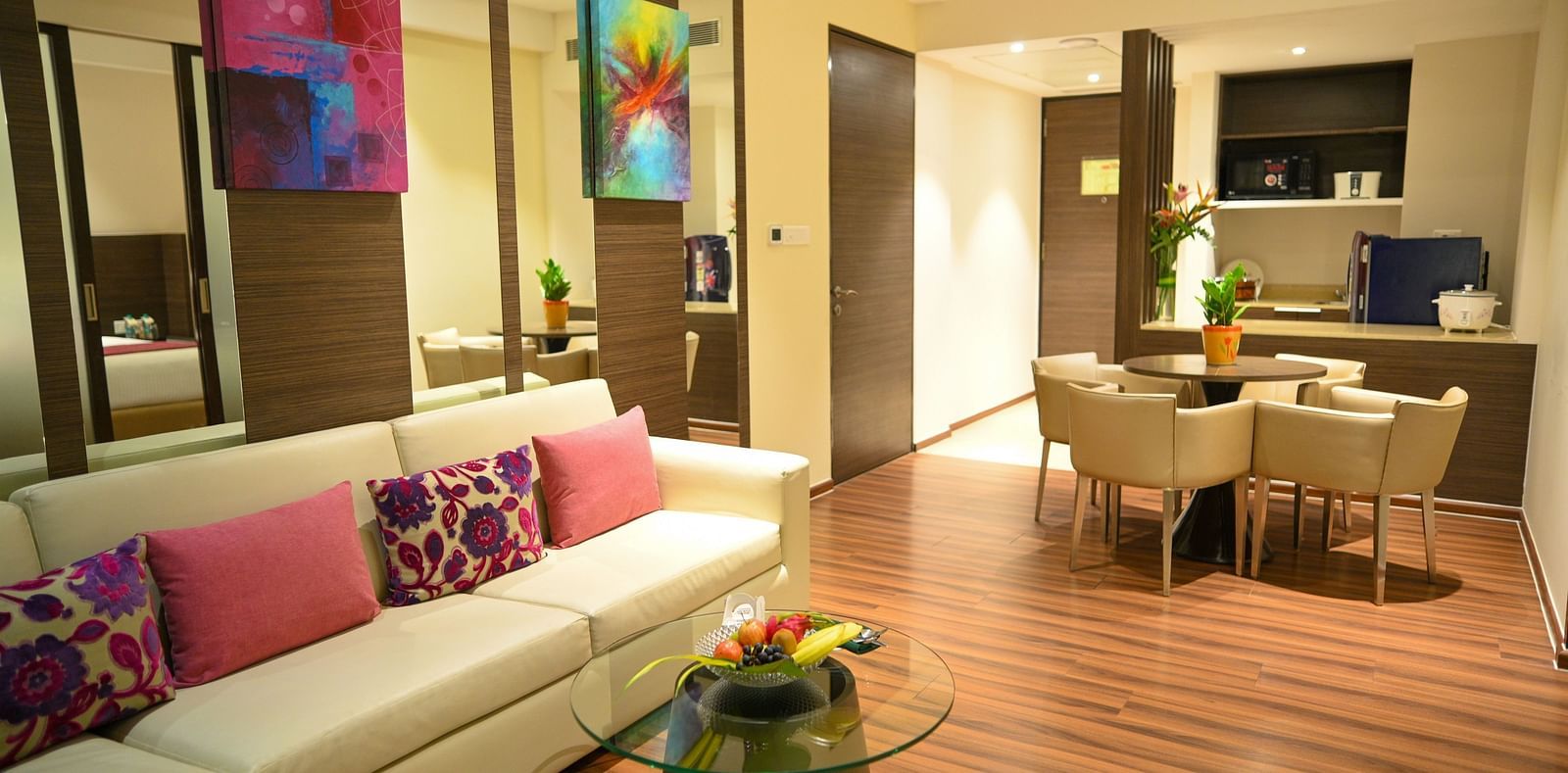 A well-lit living room with a sofa adorned with colourful pillows, a small coffee table, a dining table with chairs, and a compact kitchen.