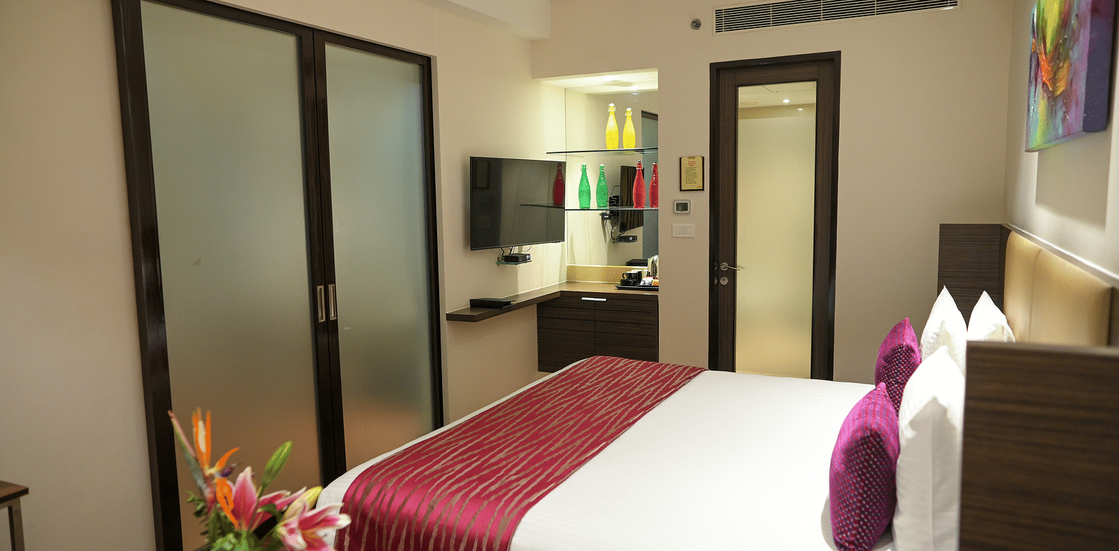 A well-lit hotel room with a king-sized bed, TV, and tea kettle. Doors lead to the bathroom and closet; a flower vase adorns a side table.