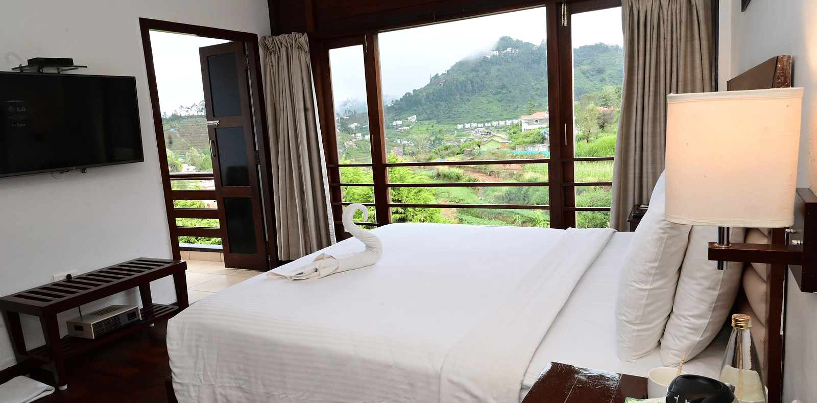 An image of a Rock Cabin Valley View with a king size bed, bed side table, lamps, flat screen TV, and an expansive window with curtains - GReaT trails Kodaikanal by GRT Hotels