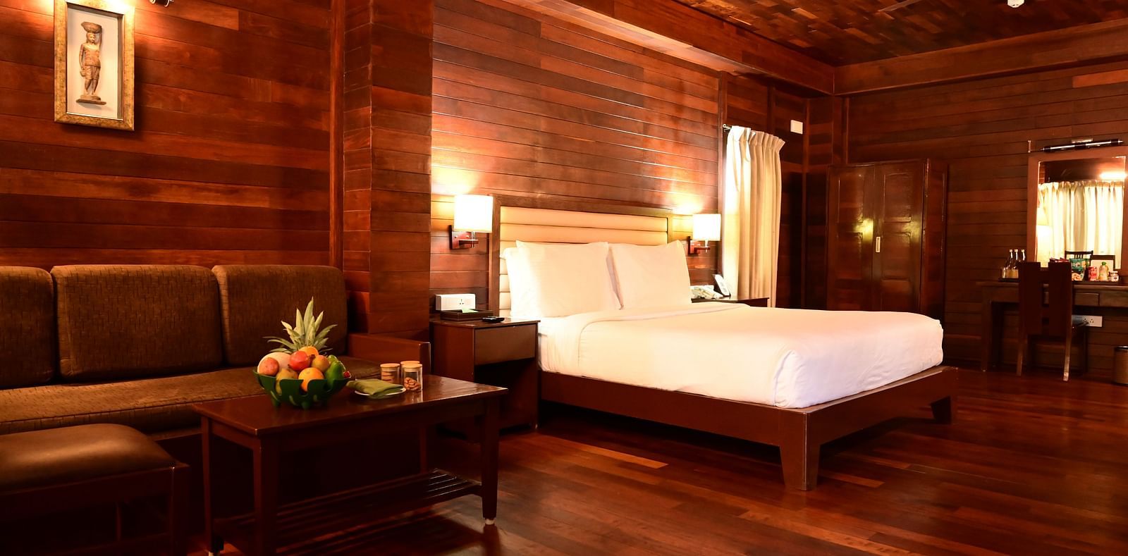 An image of a wood finish room with a king size bed, bed side table, lamps, sofa, table, modern hotel amenities, and decorative items with bright lighting - GReaT trails Kodaikanal by GRT Hotels