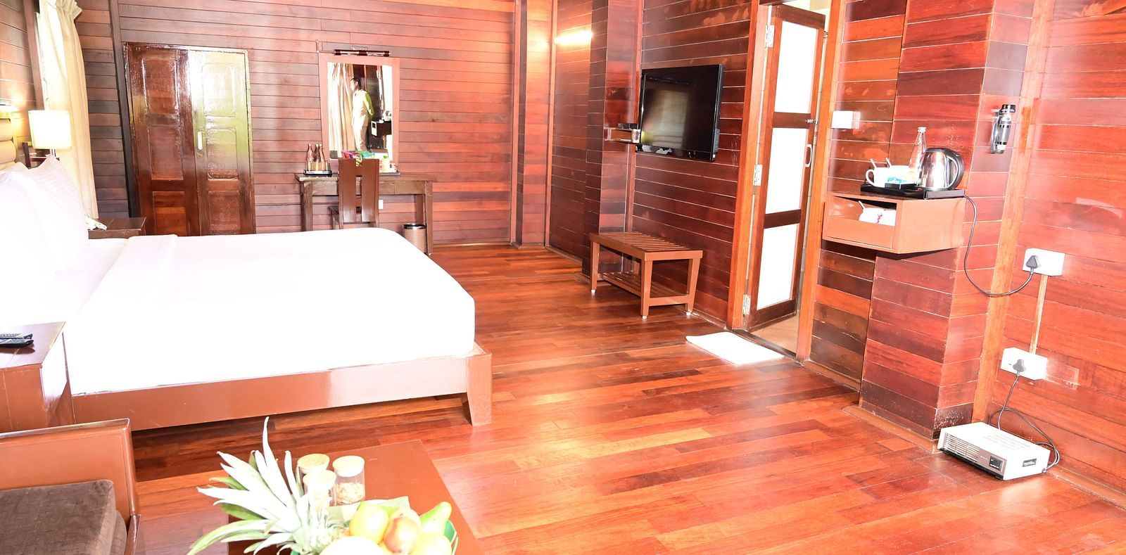 An image of a wood finish room with a king size bed, bed side table, lamps, sofa, table, fruit basket,flat screen TV, modern hotel amenities, and decorative items with bright lighting - GReaT trails Kodaikanal by GRT Hotels