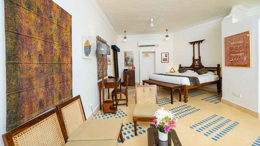 A room with a wooden four-poster bed, a seating area with beige chairs, a large abstract artwork on the wall, and blue patterned floor tiles, blending modern art with traditional decor - Tijara Fort-Palace - 19th Century, Alwar.