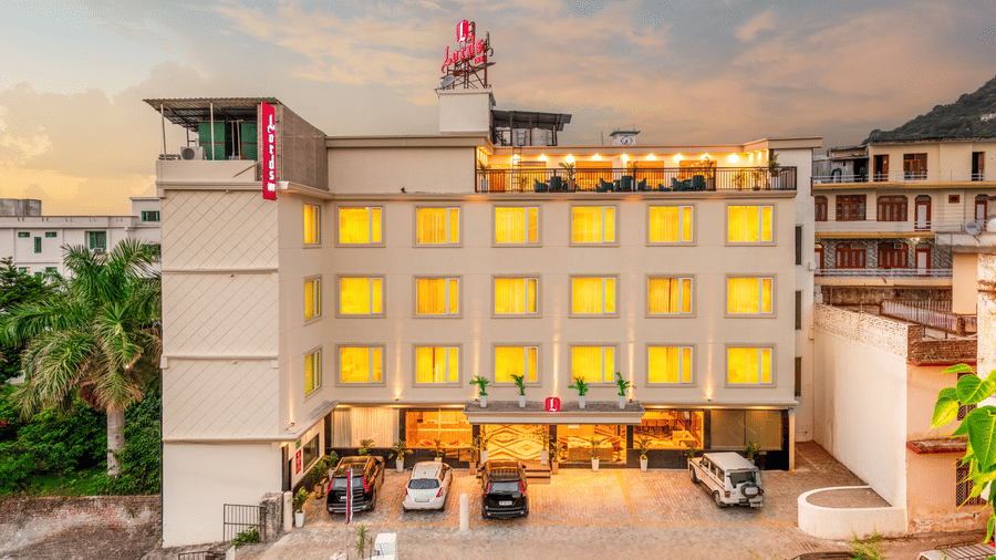 The illuminated exterior view of Lords Inn Katra shows multi storey windows, a front entrance, parked vehicles under a twilight sky.