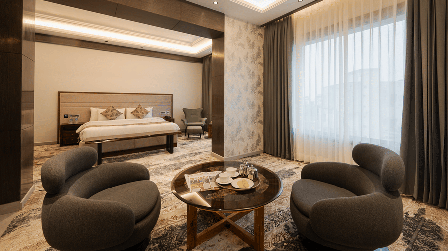 A spacious guest suite with a king-sized bed and two modern grey armchairs around a coffee table at Ananta Express, Katra.