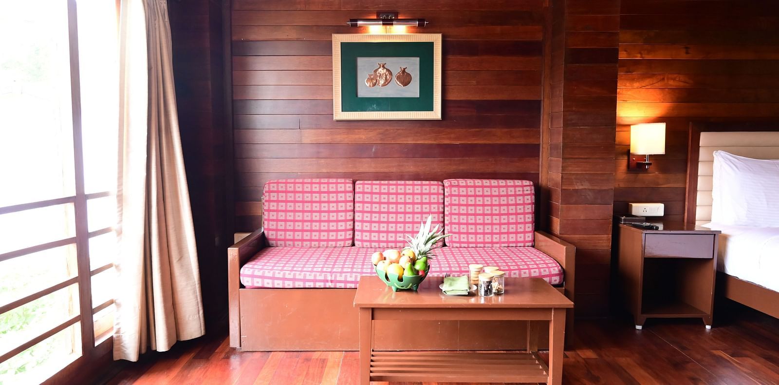 An image of a brightly lit room with a sofa, table, fruit basket, wall hangings, expansive window with curtains, and a bed - GReaT trails Kodaikanal by GRT Hotels
