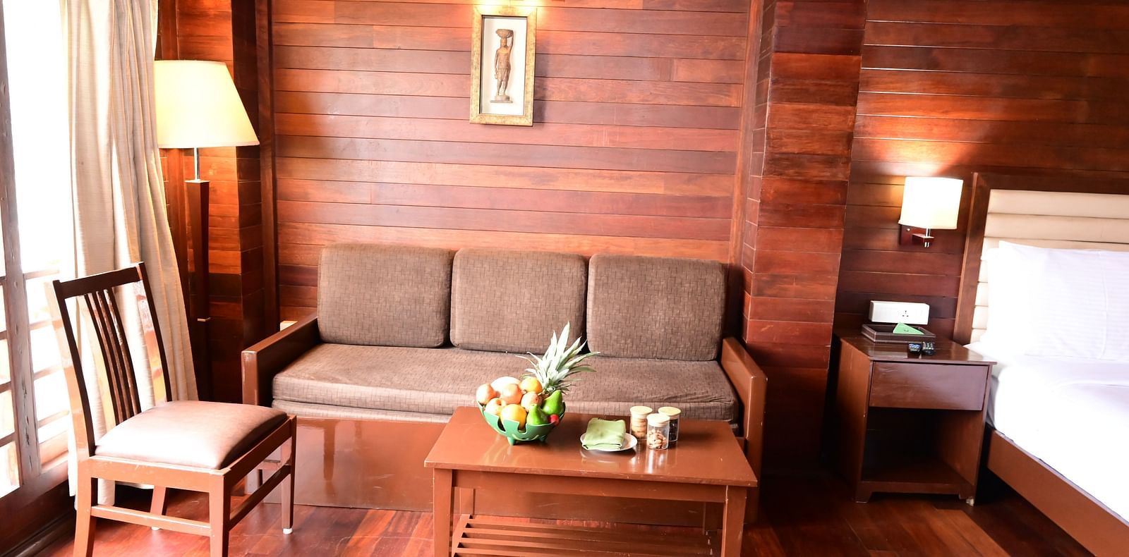 An image of a brightly lit room with a sofa, table, fruit basket, wall hangings, expansive window with curtains, and a bed - GReaT trails Kodaikanal by GRT Hotels