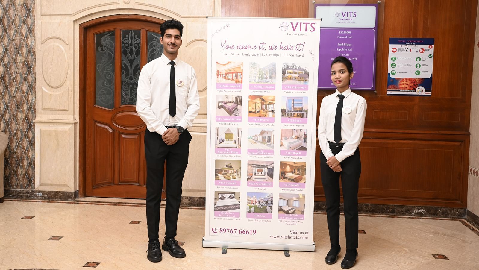 Two staff members in formal attire standing by a restaurant entrance with a hotel banner - Kamats India
