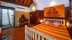 A cosy bedroom featuring a bed with striped orange and yellow linens, wood-panelled walls, an arched ceiling, and an open doorway leading to a small courtyard at Neemrana's Coconut Alley - 21st century, Tranquebar.