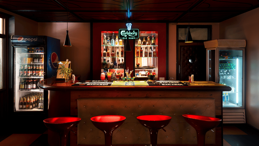 An indoor bar area, featuring a wooden counter lined with red stools, display fridges, and liquor bottles at Amantra Shilpi Resort, Udaipur.