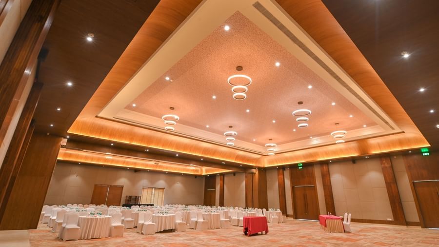  A large, brightly lit convention hall in Vedic Village Spa Resort, set up for an event, featuring rows of white chairs with gold sashes. In the foreground, two dark brown sofas with coffee tables and bottled water are visible.