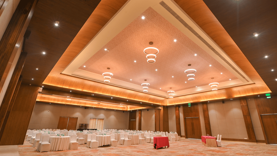 Empty banquet hall at Vedic Village Spa Resort Kolkata with warm wooden wall panelling, ornate tiered ceiling with recessed lighting, and a spacious open floor.