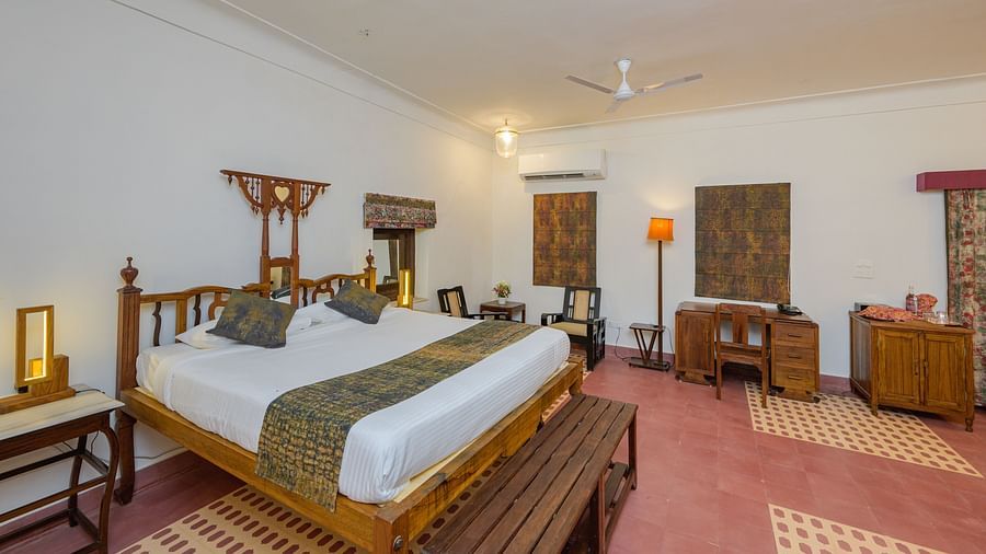 A spacious room with a wooden four-poster bed, patterned floor rugs, a dark wood desk, and an air conditioner, showcasing the room's blend of traditional and modern elements - Tijara Fort-Palace - 19th Century, Alwar.