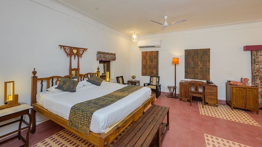 A spacious room with a wooden four-poster bed, patterned floor rugs, a dark wood desk, and an air conditioner, showcasing the room's blend of traditional and modern elements - Tijara Fort-Palace - 19th Century, Alwar.