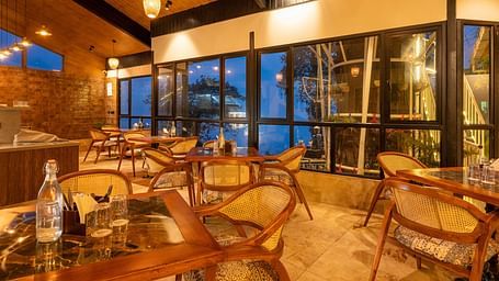 An elegant restaurant interior at dusk with large windows offering a view of the outside, warm lighting, and wooden furniture creating a cosy dining atmosphere- Arcadia Heritage Resort , Darjeeling