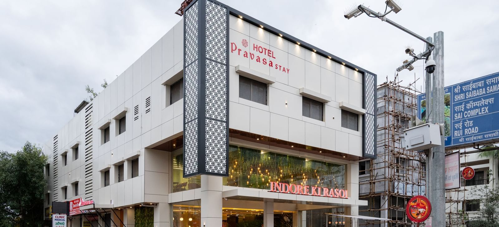 The exterior of Pravasa Stay, Shirdi is a three-storey building with large storefront windows at the ground level.