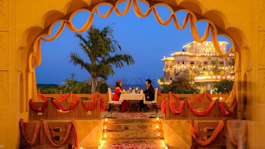 A romantic dinner setup under an arched doorway decorated with orange fabric, overlooking the illuminated fort-palace at dusk, featuring two people seated at a table - Tijara Fort-Palace - 19th Century, Alwar.