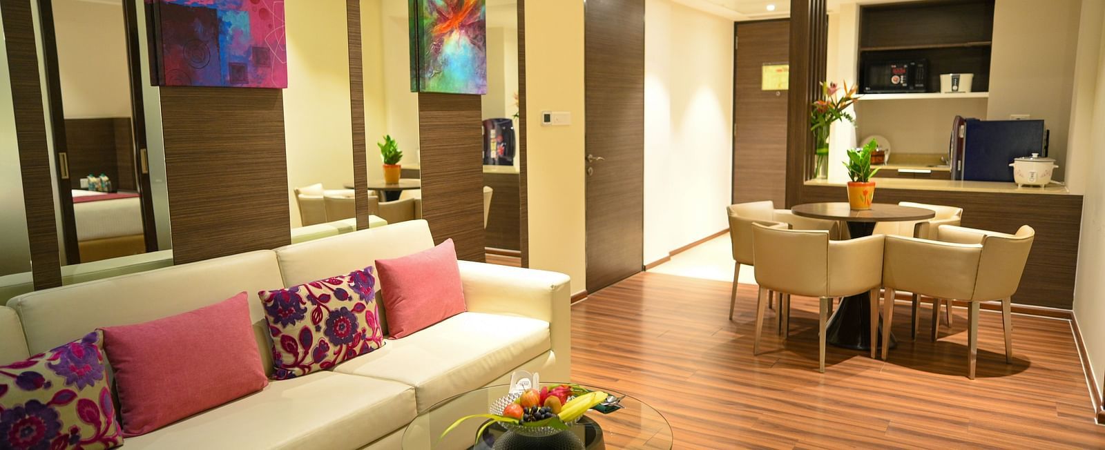 A well-lit living room with a sofa adorned with colourful pillows, a small coffee table, a dining table with chairs, and a compact kitchen.