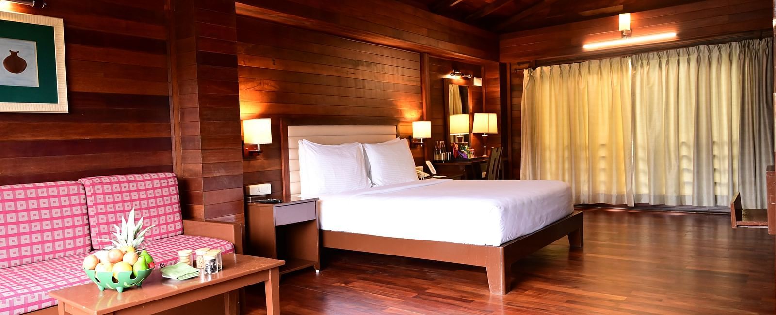 An image of a wood finish room with a king size bed, bed side table, lamps, sofa, table, and decorative items with bright lighting - GReaT trails Kodaikanal by GRT Hotels