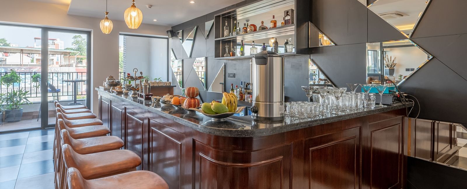 A dimly lit bar area at Vishranti Hotel, Dehradun shows a dark wooden bar counter with stools, a selection of beverages, and a mirrored wall with geometric patterns. 