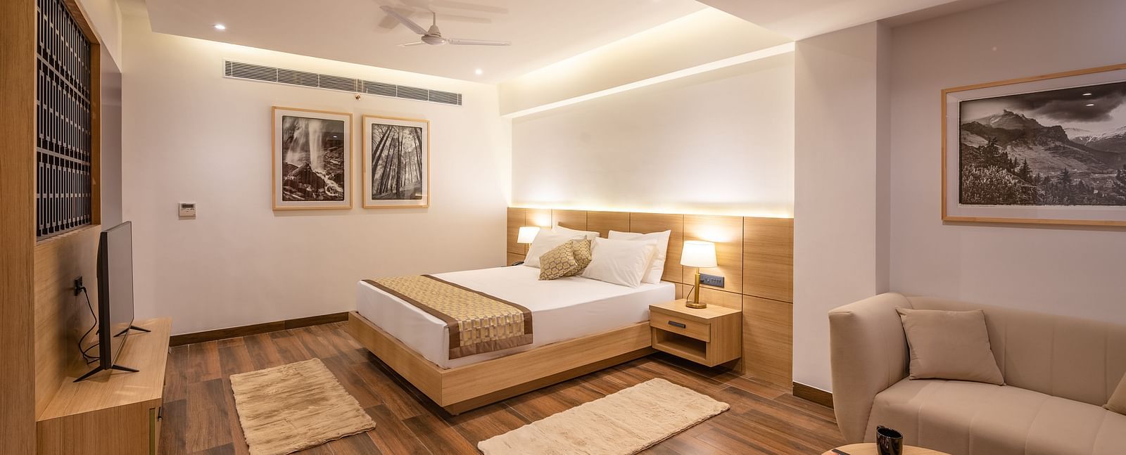 Side shot of Suite at Vishranti Hotel, Dehradun featuring a king bed with side tables, a sofa, a TV, large window with curtains and bed lamps. 