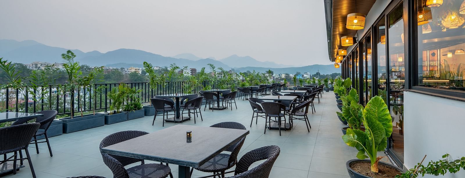 Side shot of an outdoor seating area at Vishranti Hotel, Dehradun with a seating area features multiple tables with chairs, and plants are visible in the background.