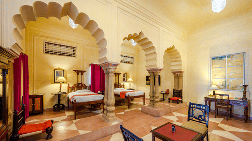 A room with two beds, two tables, chairs, and an arched entryway in Tijara Fort-Palace - 19th Century, Alwar.