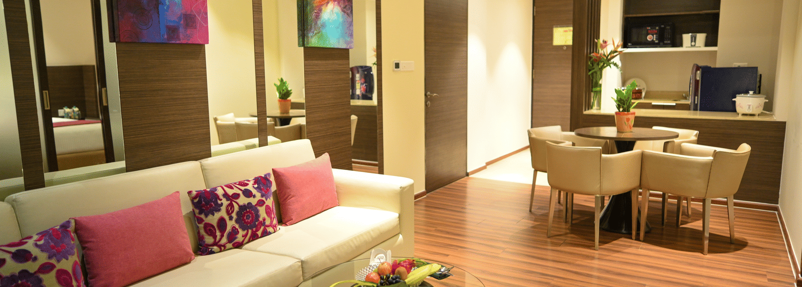 A well-lit living room with a sofa adorned with colourful pillows, a small coffee table, a dining table with chairs, and a compact kitchen.