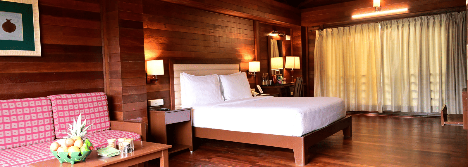 An image of a wood finish room with a king size bed, bed side table, lamps, sofa, table, and decorative items with bright lighting - GReaT trails Kodaikanal by GRT Hotels