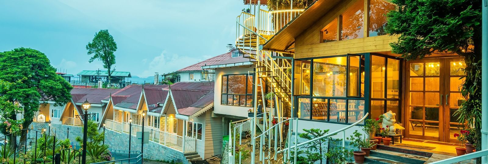 A close up view of a cosy hillside establishment glowing warmly at twilight, with its wooden architecture and inviting lights reflecting a welcoming ambience amidst lush greenery- Arcadia Heritage resort, Darjeeling