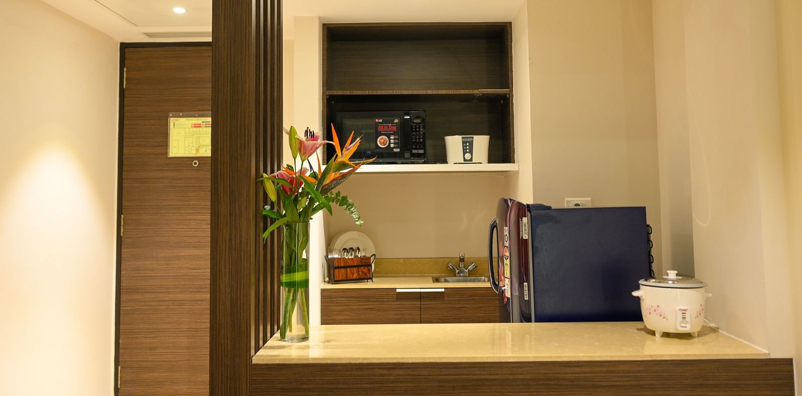 A compact kitchen near the entry gate, equipped with a refrigerator, microwave, cutlery, toaster, electric cooker, sink, flower vase, and countertop.