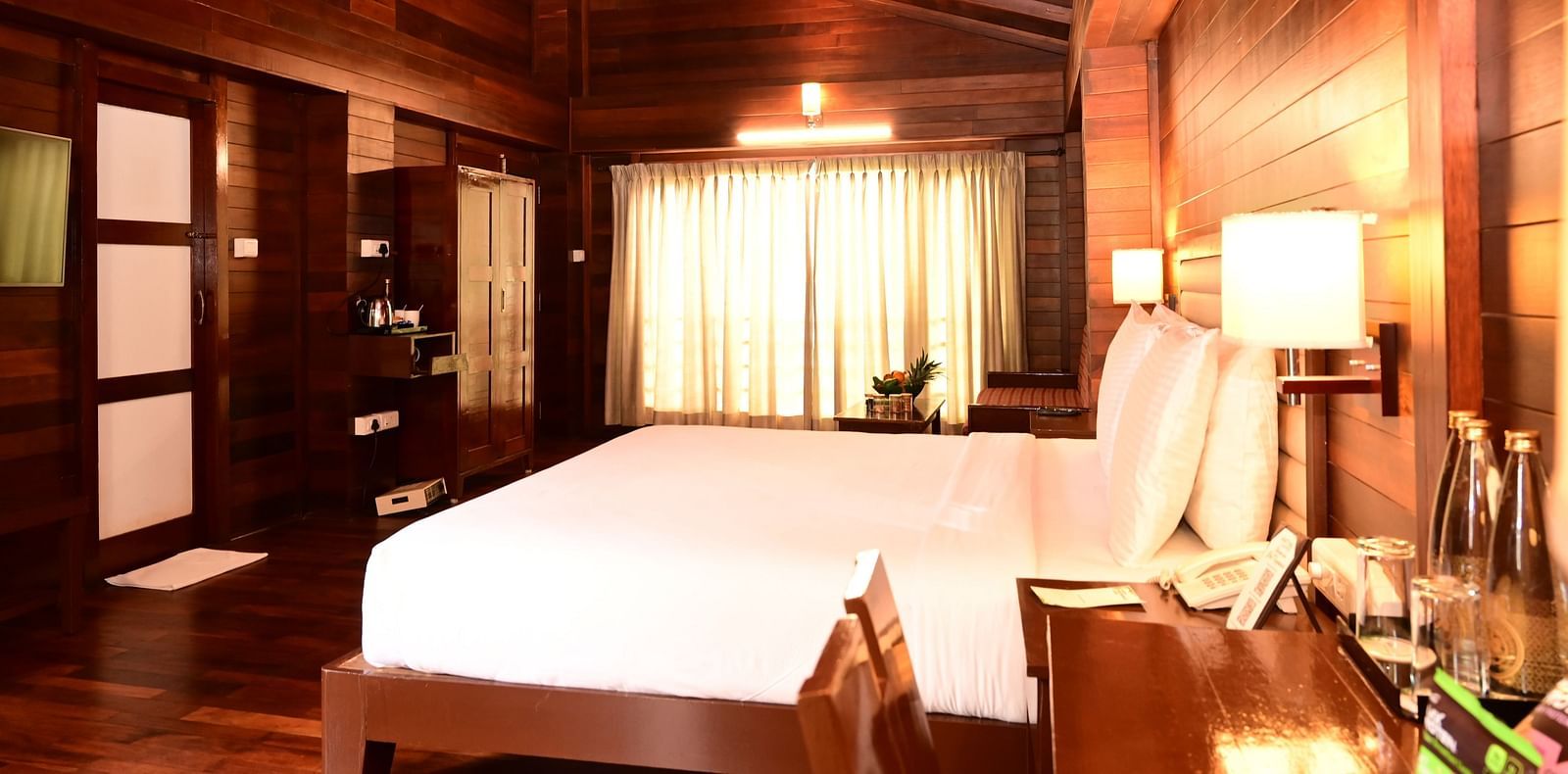 An image of a wood finish room with a king size bed, bed side table, lamps, sofa, table, flat screen TV, and decorative items with bright lighting - GReaT trails Kodaikanal by GRT Hotels