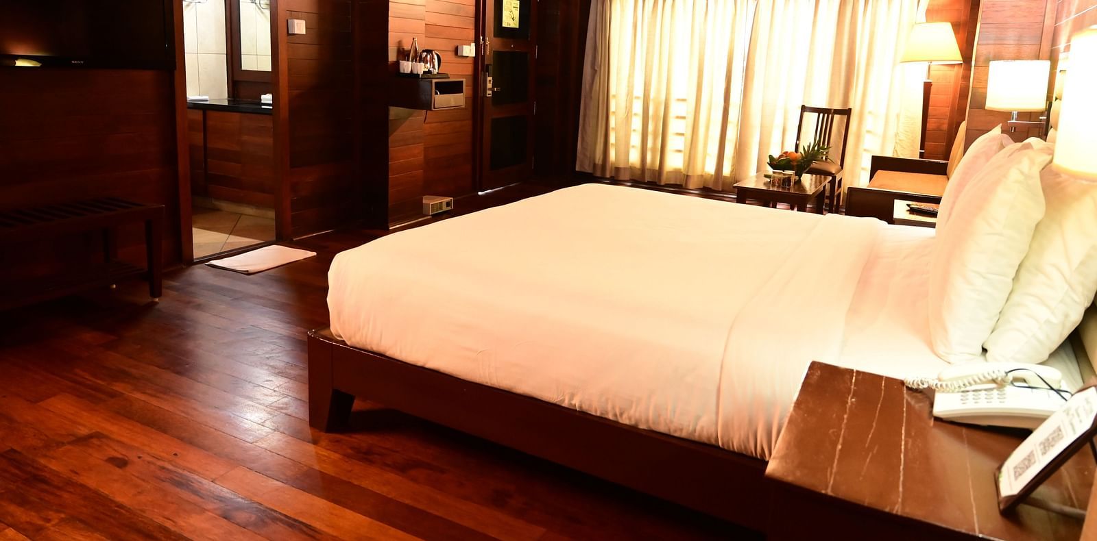 An image of a wood finish room with a king size bed, bed side table, lamps, sofa, table, flat screen TV, and decorative items with bright lighting - GReaT trails Kodaikanal by GRT Hotels