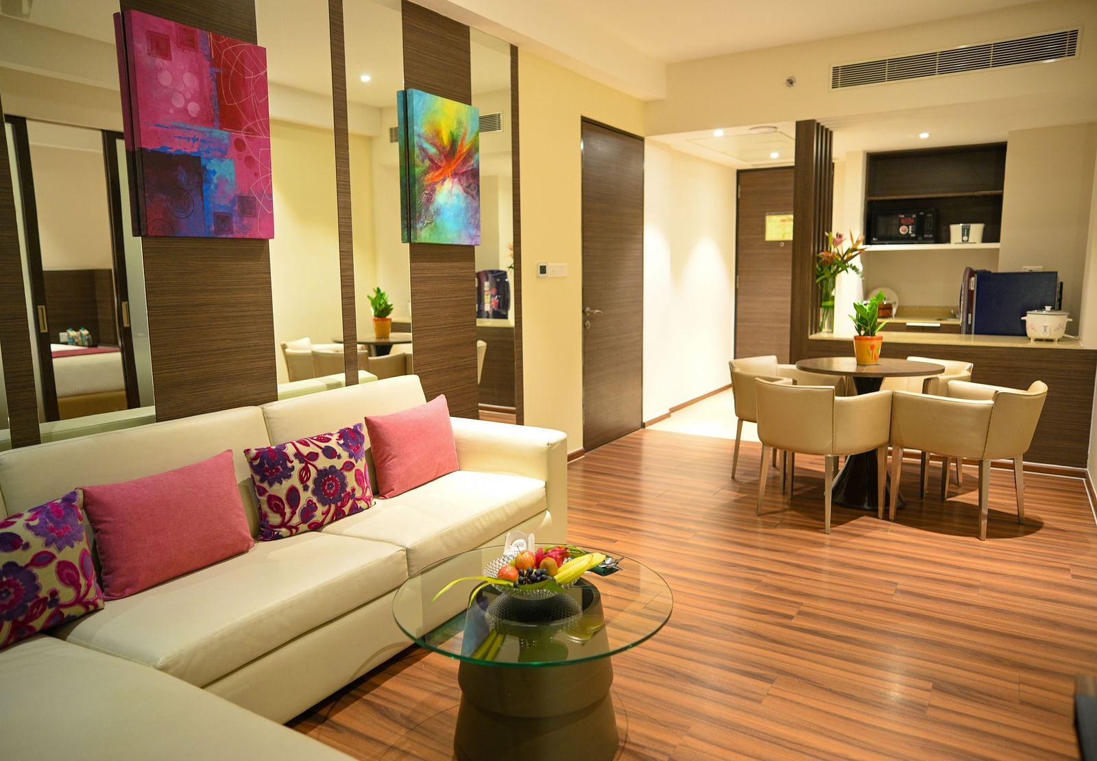 A well-lit living room with a sofa adorned with colourful pillows, a small coffee table, a dining table with chairs, and a compact kitchen.