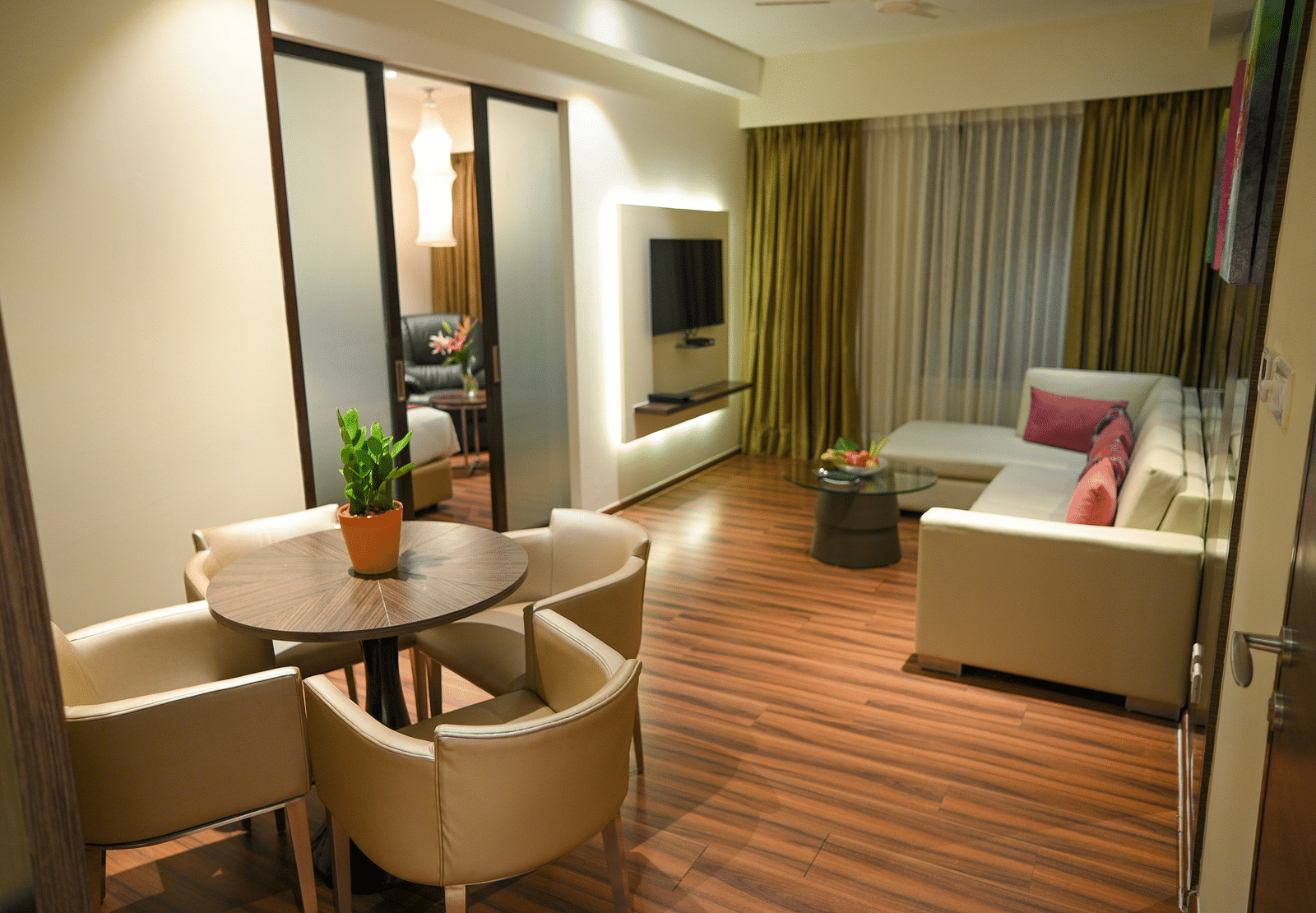 A spacious suite with a living area, off-white couch, TV, dining table, and a sliding door to the bedroom with a bed and side table — perfect for an apartment stay in Kakinada.