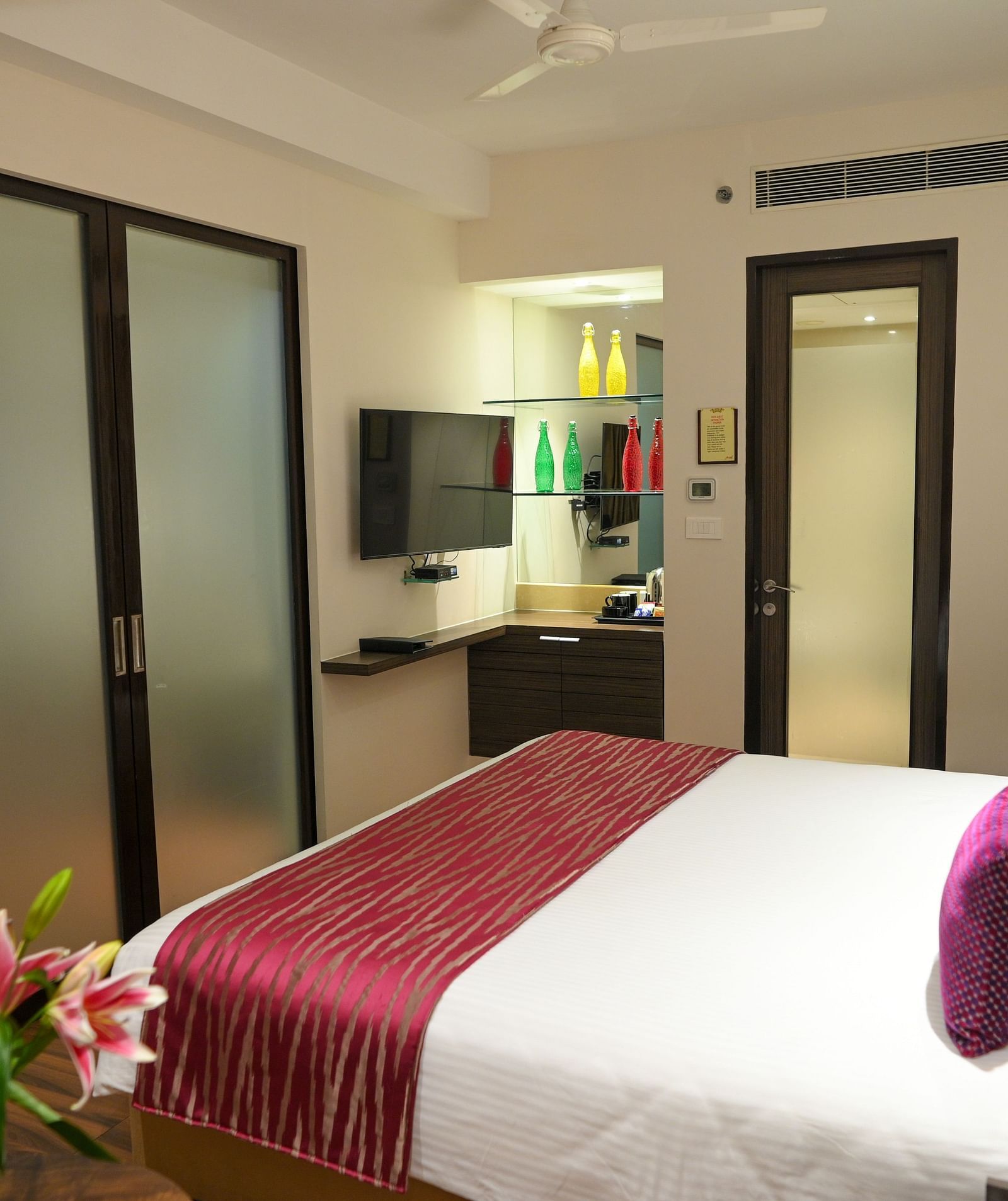 A well-lit hotel room with a king-sized bed, TV, and tea kettle. Doors lead to the bathroom and closet; a flower vase adorns a side table.
