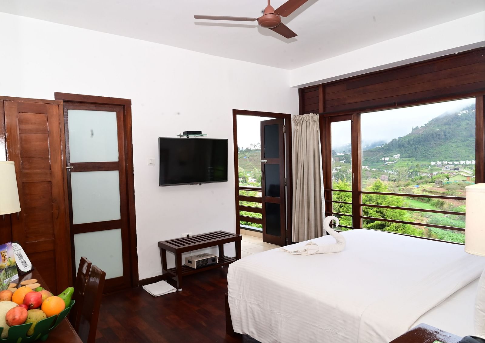 An image of a Rock Cabin Valley View with a king size bed, bed side table, lamps, flat screen TV, and an expansive window with curtains - GReaT trails Kodaikanal by GRT Hotels