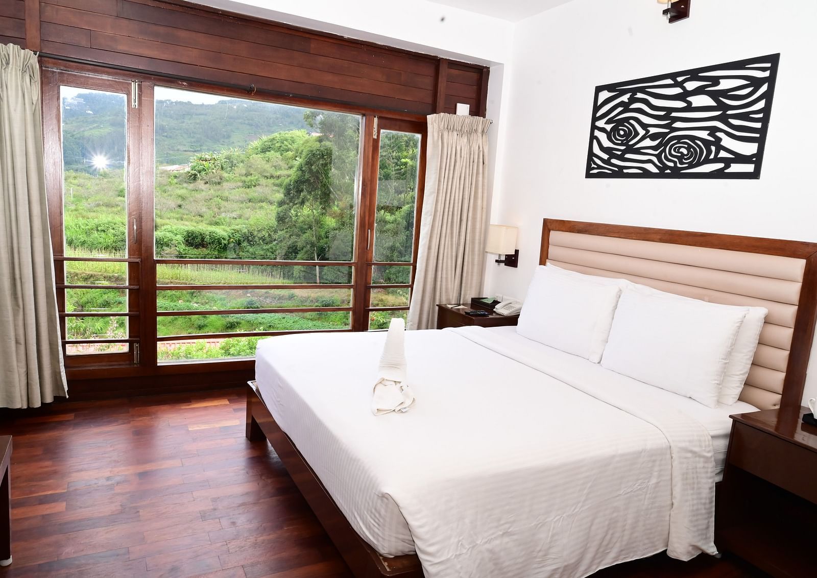 An image of a Rock Cabin Valley View with a king size bed, bed side table, lamps, wall paintings, and an expansive window with curtains - GReaT trails Kodaikanal by GRT Hotels