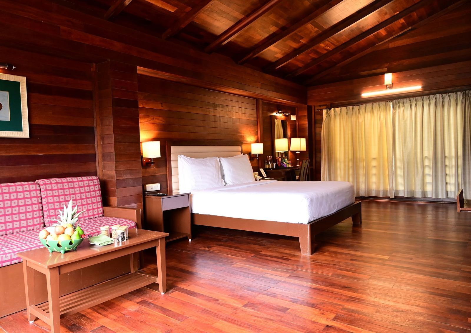 An image of a wood finish room with a king size bed, bed side table, lamps, sofa, table, and decorative items with bright lighting - GReaT trails Kodaikanal by GRT Hotels