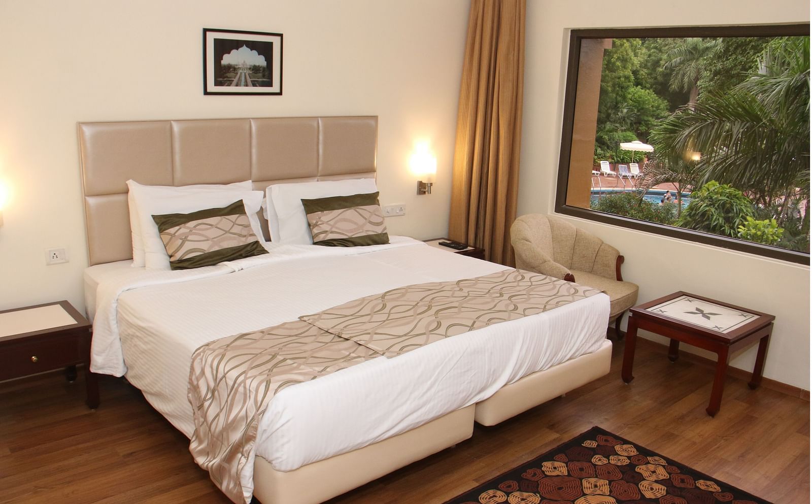 A room with a large bed, beige headboard, wooden flooring, bedside tables, and a window overlooking greenery and a swimming pool - Hotel Clarks Shiraz, Agra