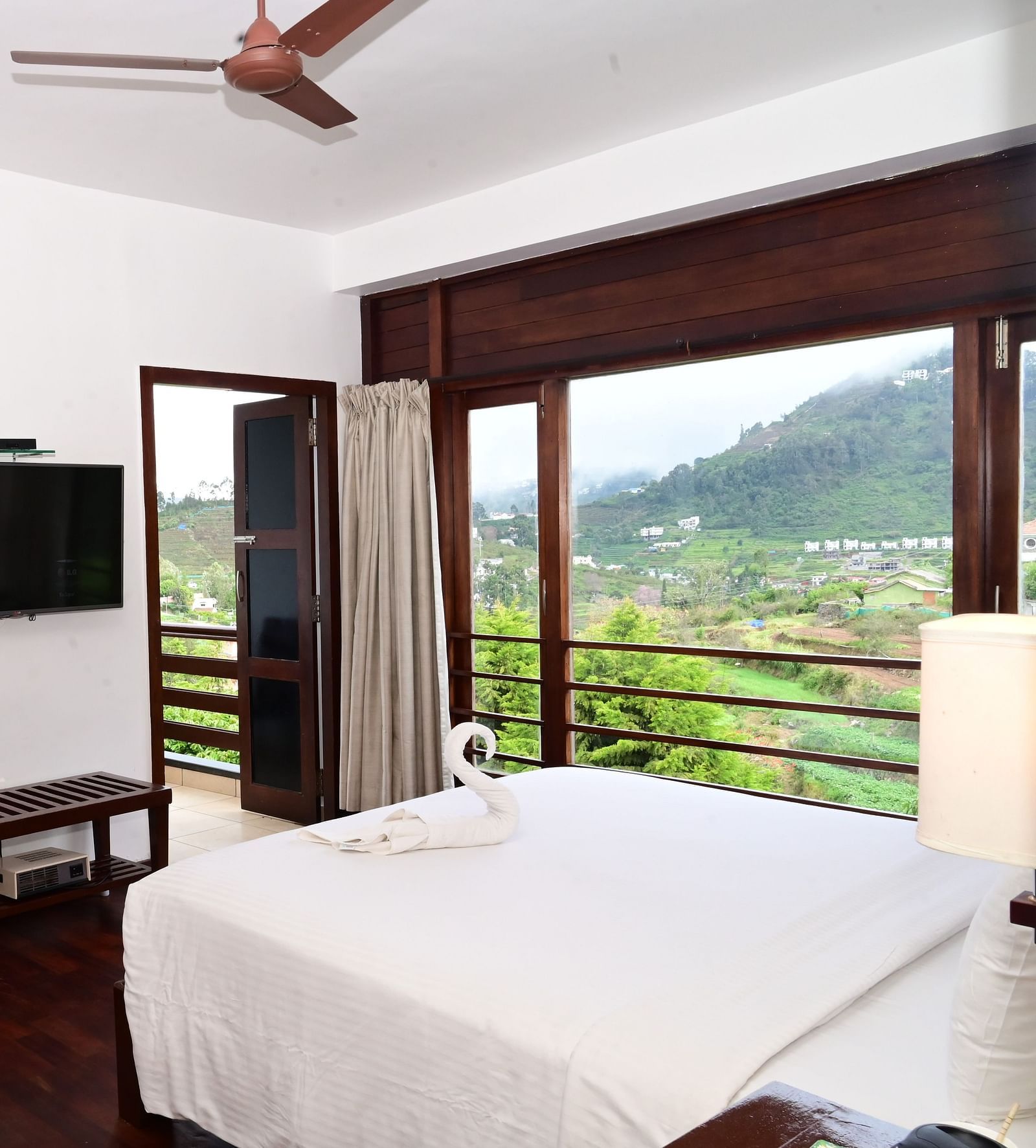 An image of a Rock Cabin Valley View with a king size bed, bed side table, lamps, flat screen TV, and an expansive window with curtains - GReaT trails Kodaikanal by GRT Hotels